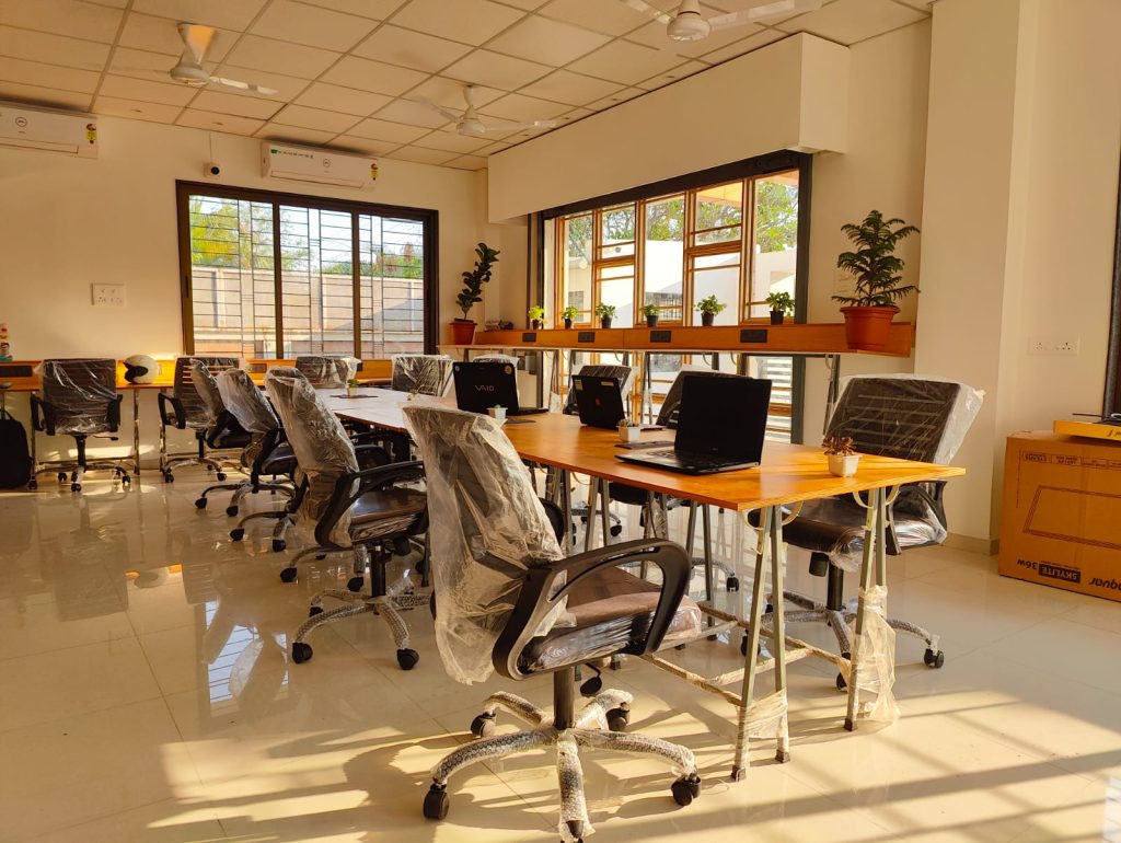 The image shows a well-lit office space with large windows letting in plenty of natural light. There are multiple workstations with wooden desks arranged in a row, each equipped with a black office chair and a laptop. The chairs and some equipment are covered with transparent plastic, suggesting either new installations or an intent to keep them dust-free. Plants are placed at intervals along the window sill, adding a touch of greenery. On the far right, there's a tall plant in a pot, and cardboard boxes are stacked against the wall, possibly containing office supplies or equipment. The floor reflects the sunlight, indicating it is polished and shiny.